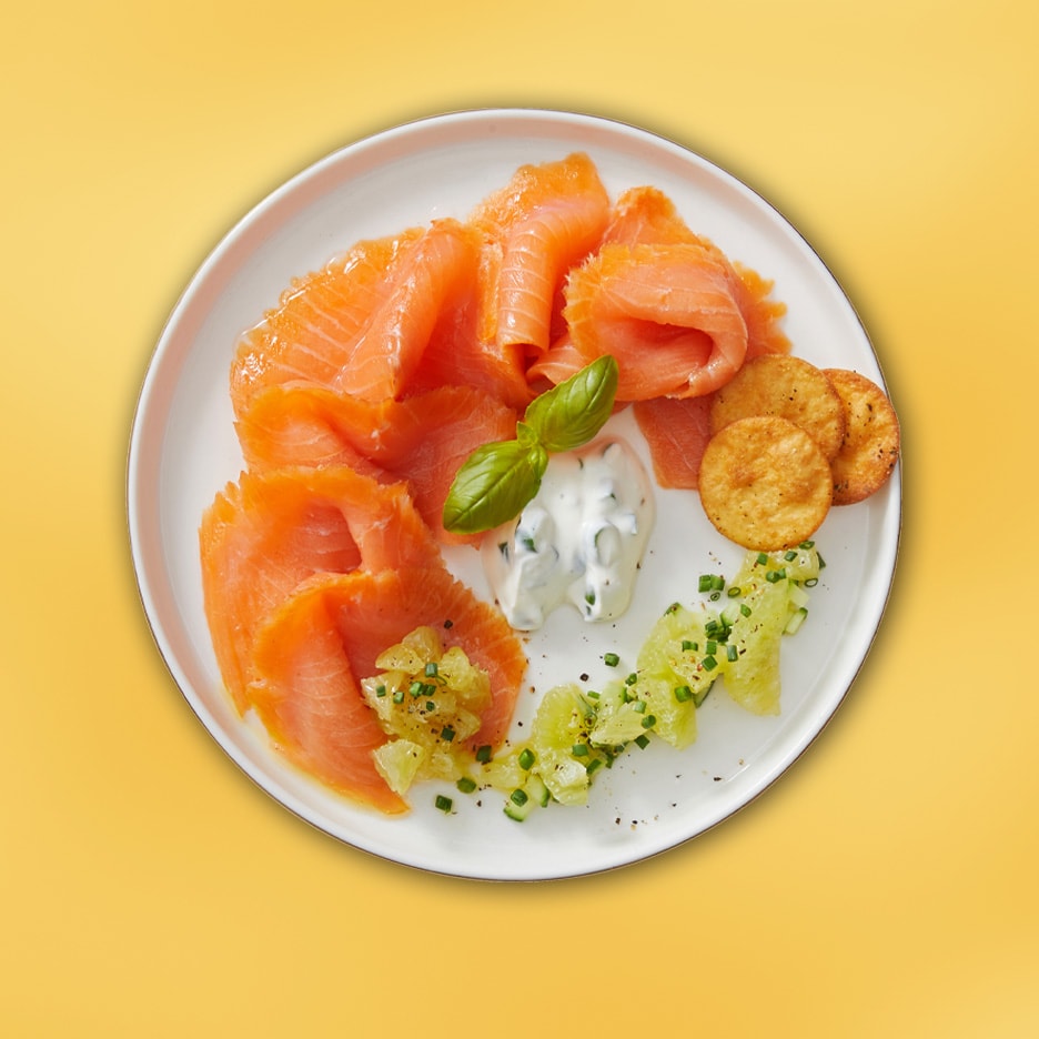 Røget laks med dildcreme, kiks og agurkesalat på en hvid tallerken.