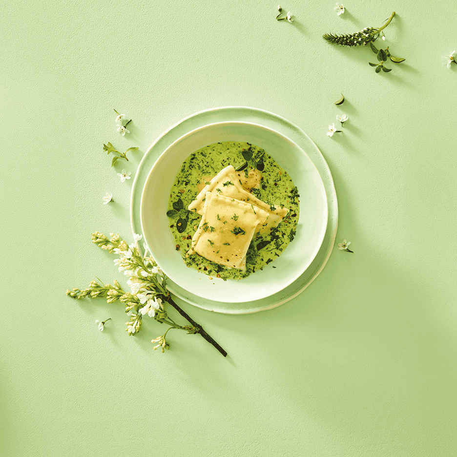 Ravioli i grøn sauce med krydderurter og blomster på en lysegrøn baggrund.