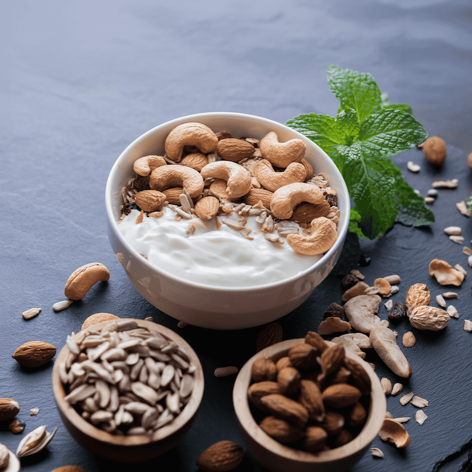 Skål med yoghurt, cashewnødder, mandler og solsikkekerner på en mørk overflade.