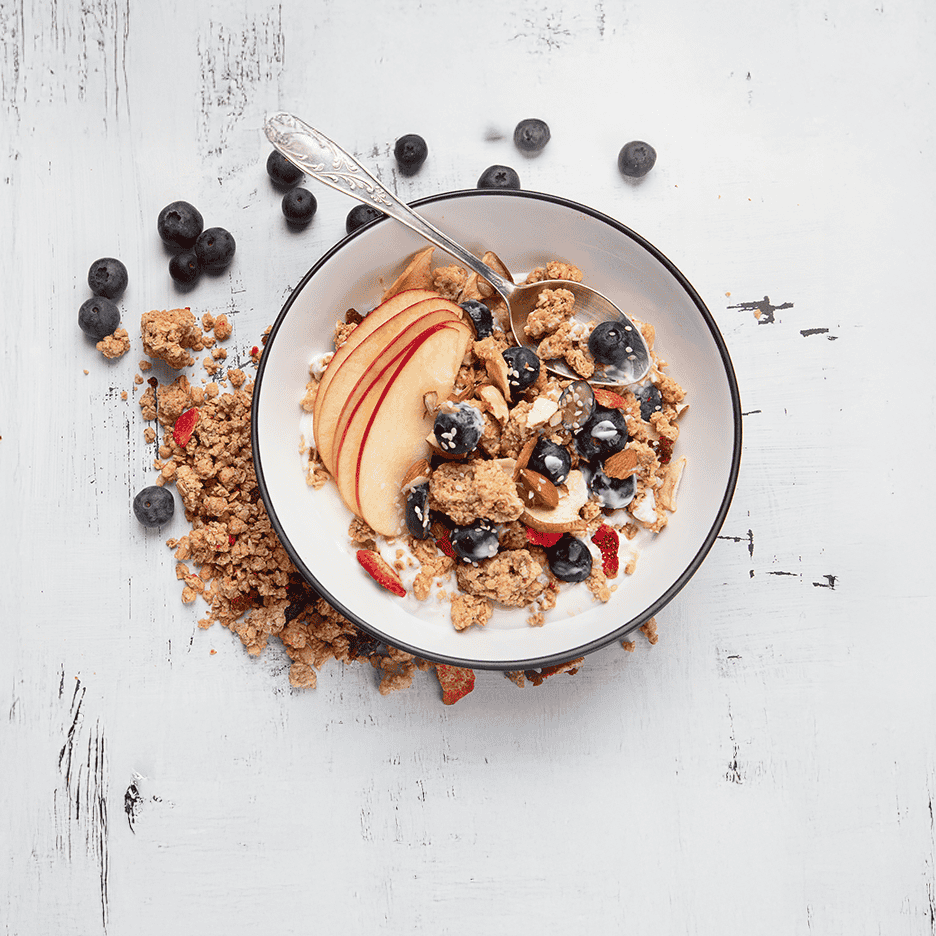 Skål med granola, yoghurt, æble, blåbær og mandler på et hvidt træbord.