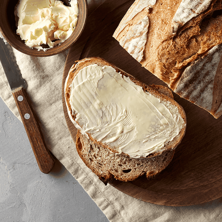 Skive rugbrød med smør, en skål smør og et brød på et træbræt.
