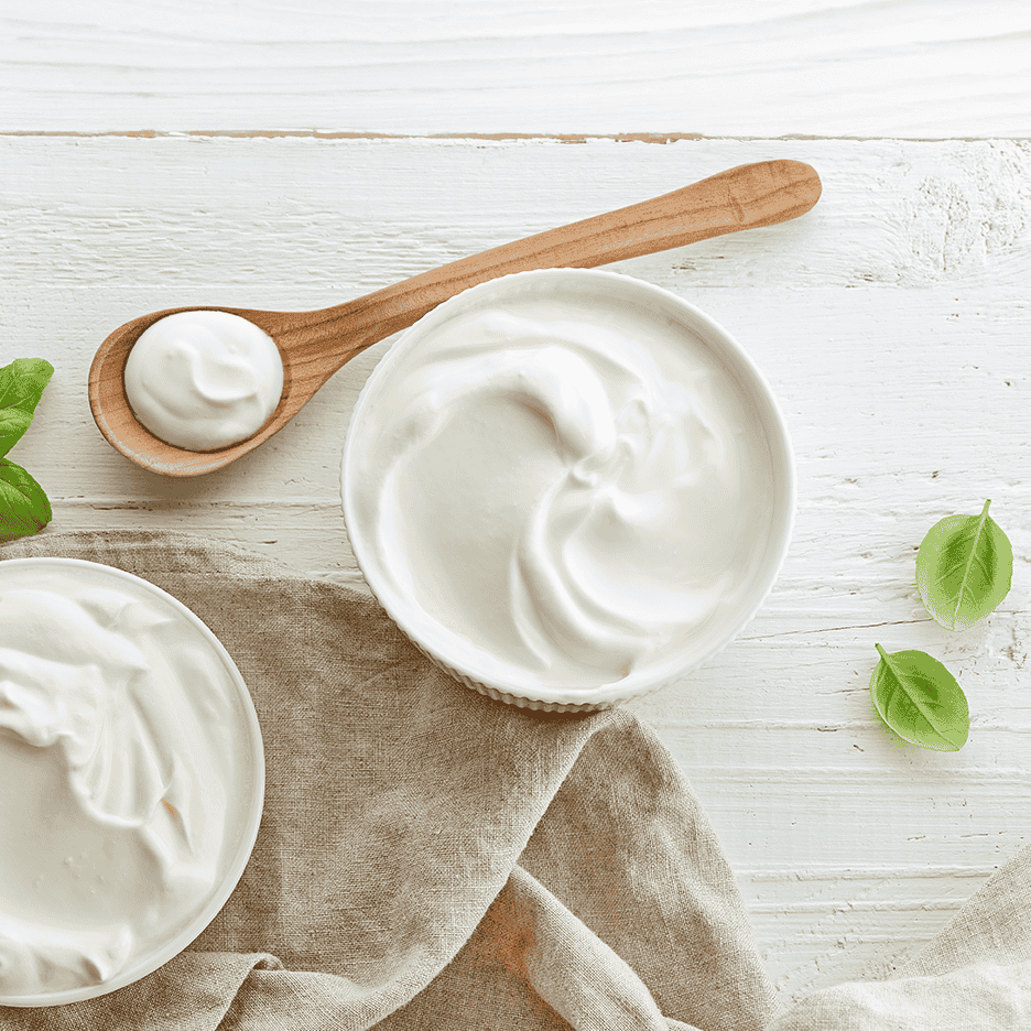 To skåle og en ske med hvid creme fraiche eller yoghurt på et hvidt træbord med basilikumblade.