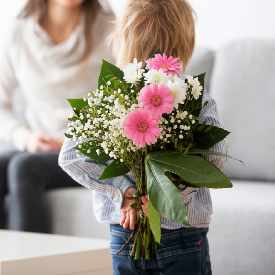 Barn holder en buket blomster med lyserøde gerbera og hvide blomster bag ryggen til en kvinde.