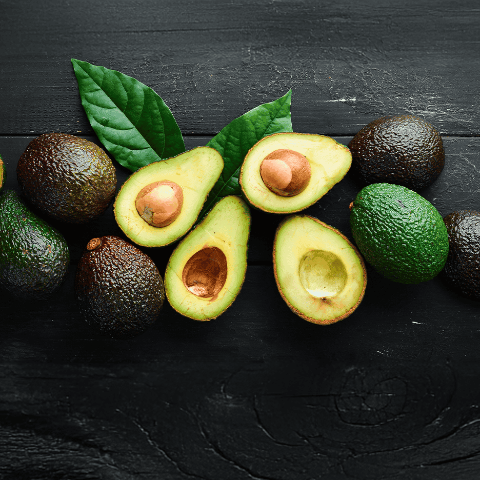 Friske hele og halverede avocadoer med blade på et mørkt træunderlag.