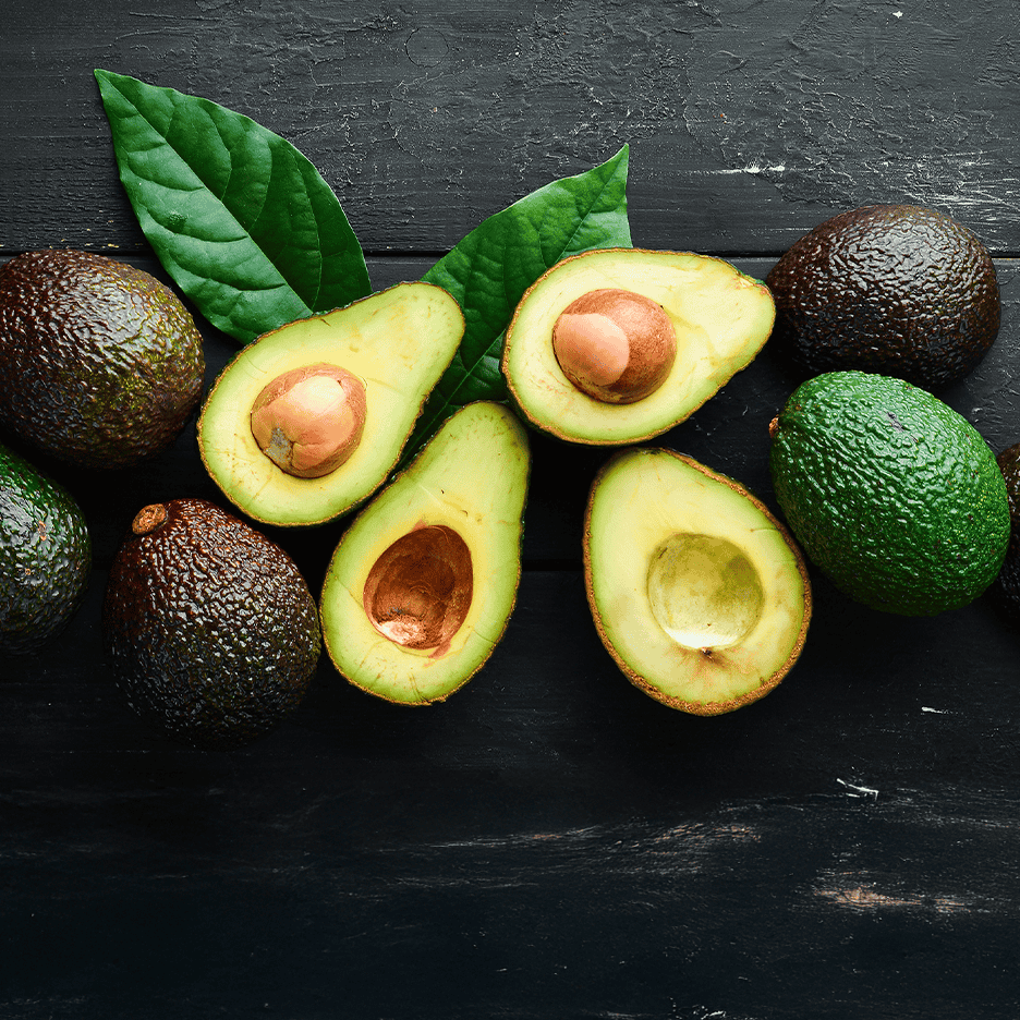 Friske hele og halverede avocadoer med blade på et mørkt træunderlag.