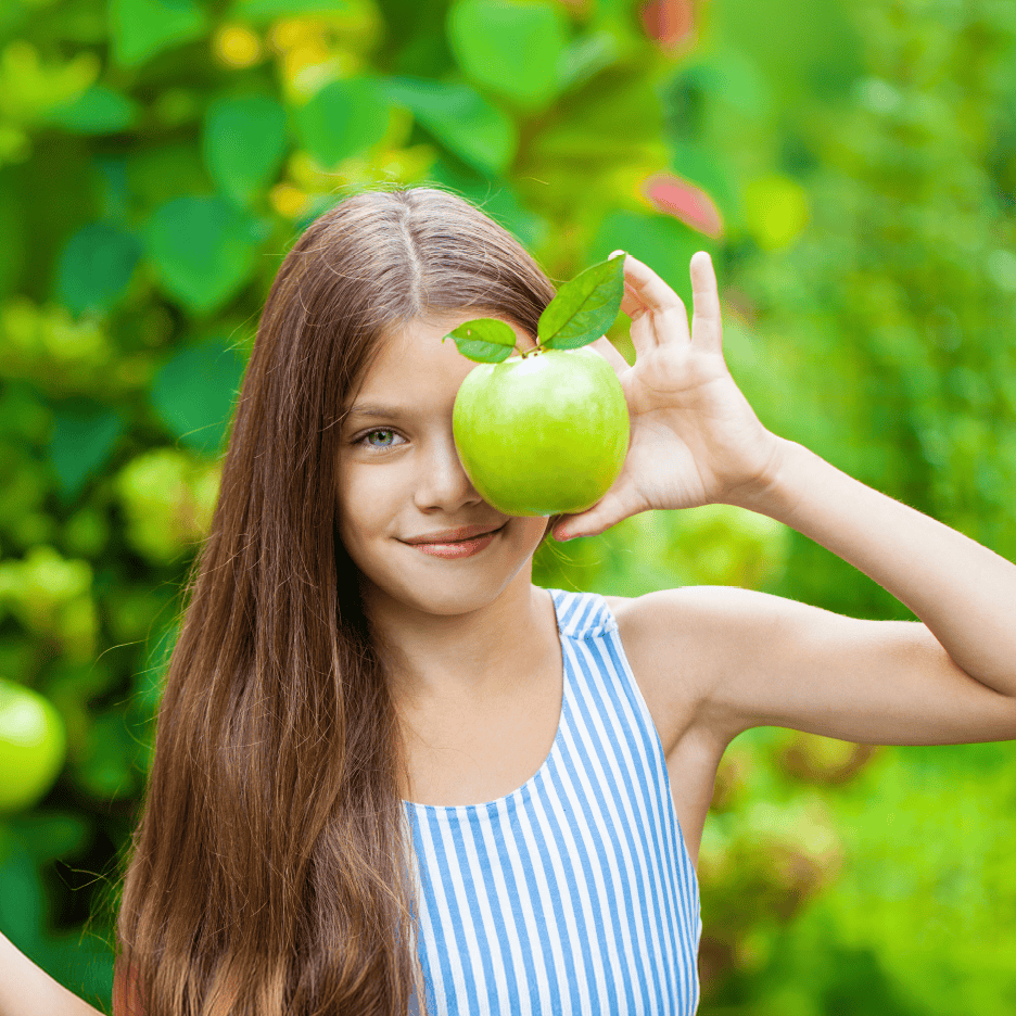 Pige med langt hår holder et grønt æble foran sit øje, smilende mod kameraet.