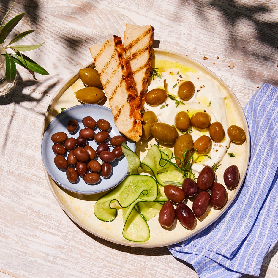 En tallerken med oliven, grillet brød, agurkeskiver og en hvid dip.