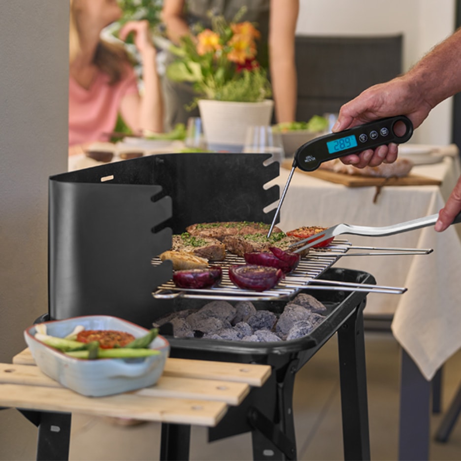 En mand griller kød og grøntsager på en kulgrill og bruger et digitalt termometer.