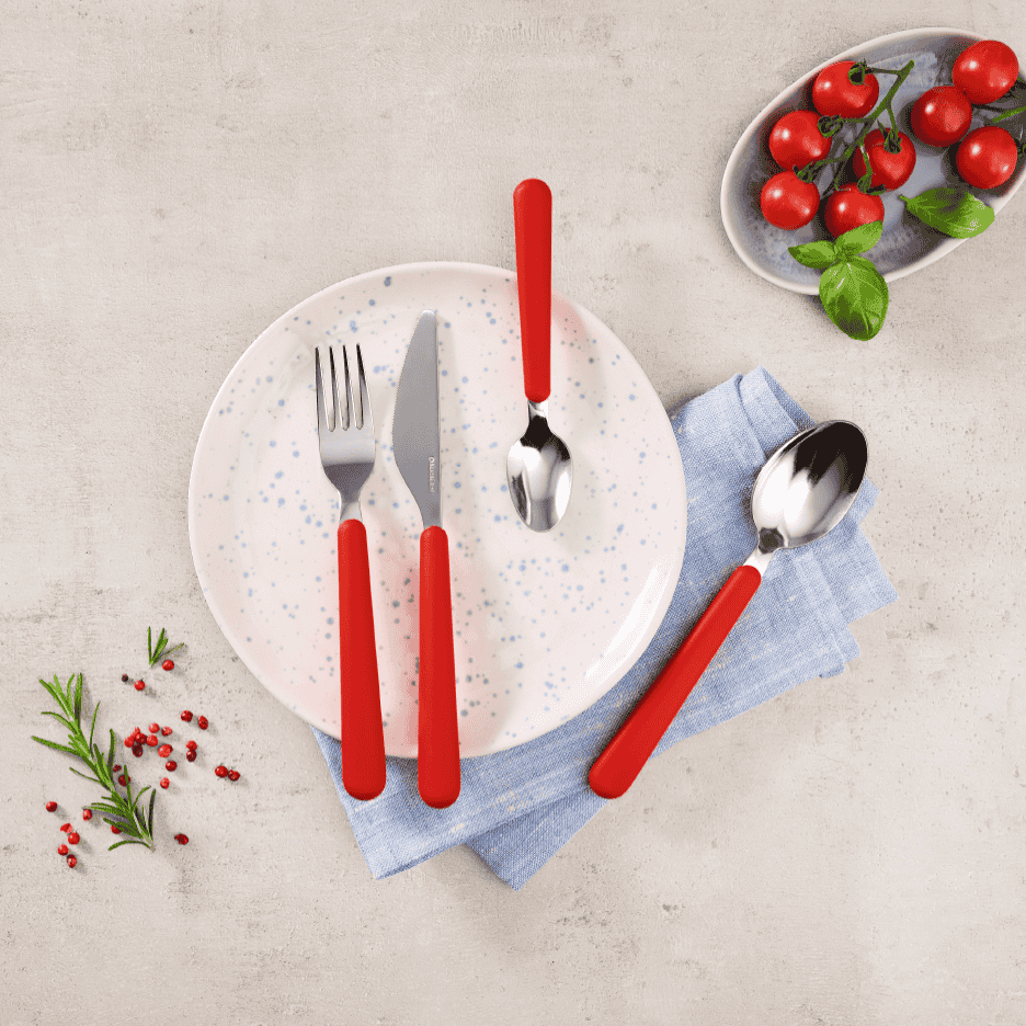 Rødt bestiksæt med tallerken, serviet, tomater og krydderurter på et lyst underlag.
