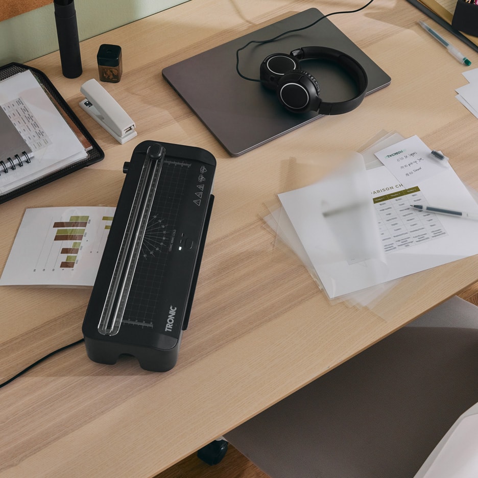 Kontorbord med lamineringsmaskine, bærbar computer, hovedtelefoner og papirer.