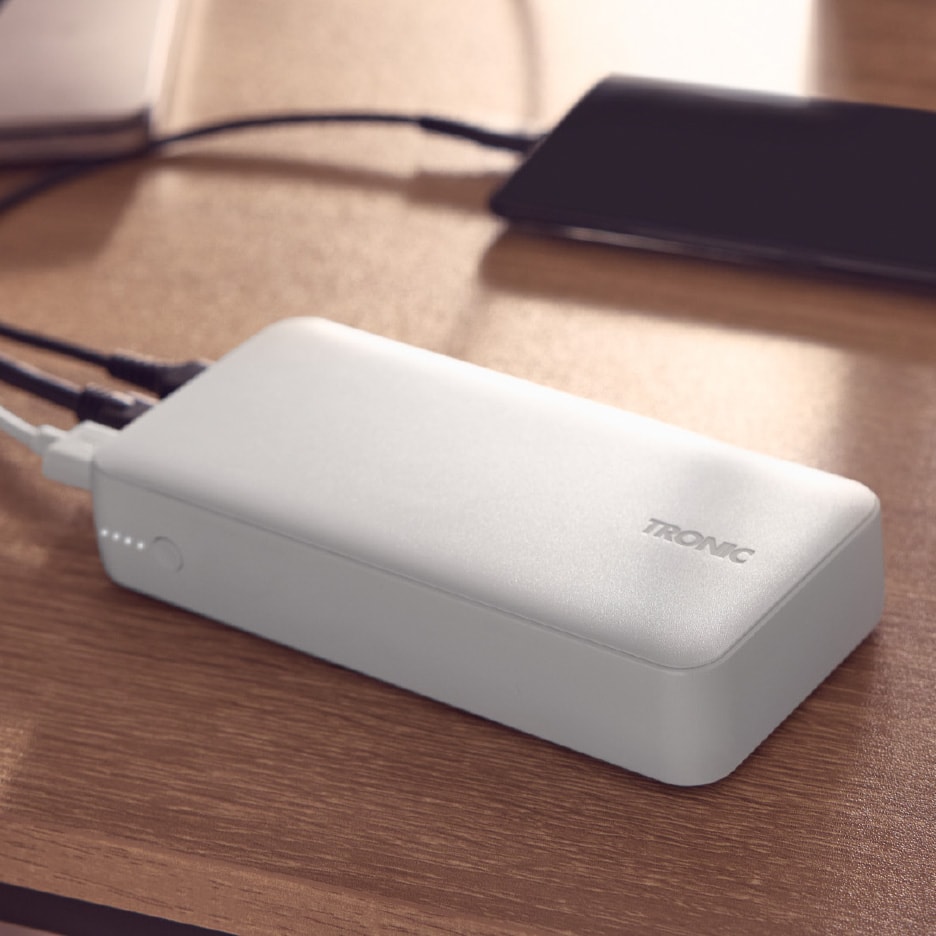Hvid powerbank med tilsluttede kabler og en smartphone på et træbord.