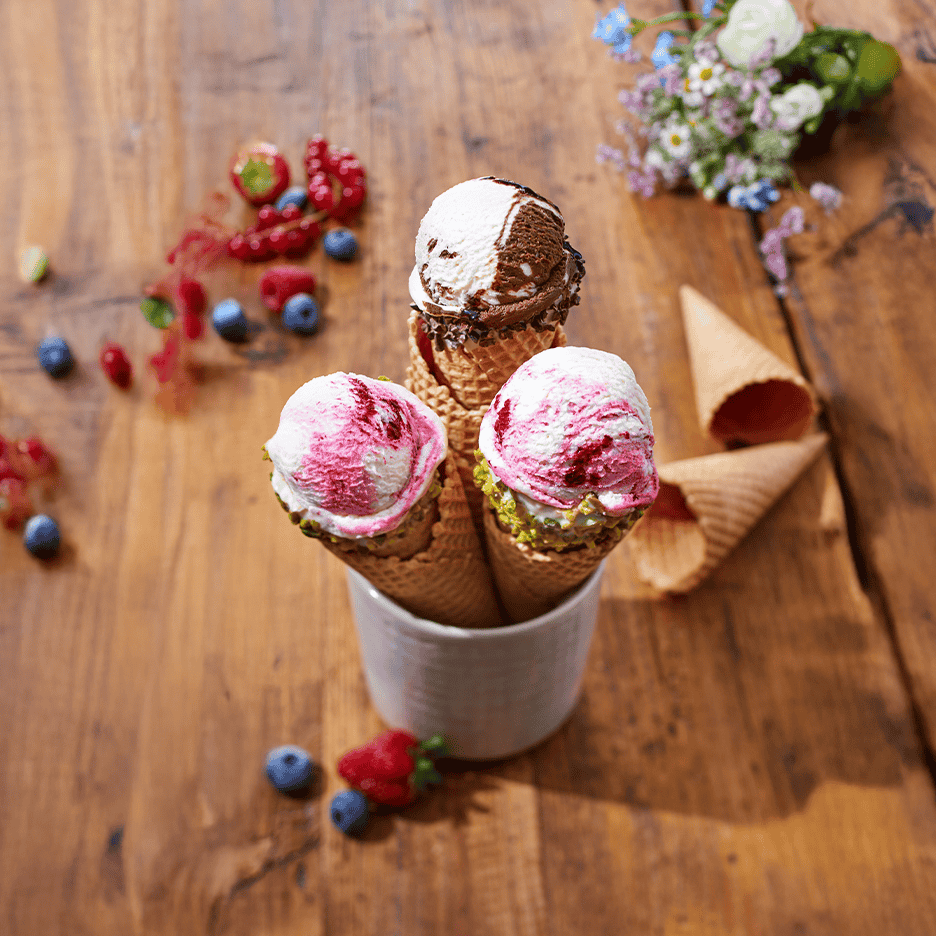 Tre isvafler med forskellige smagsvarianter i en kop, omgivet af bær og blomster.