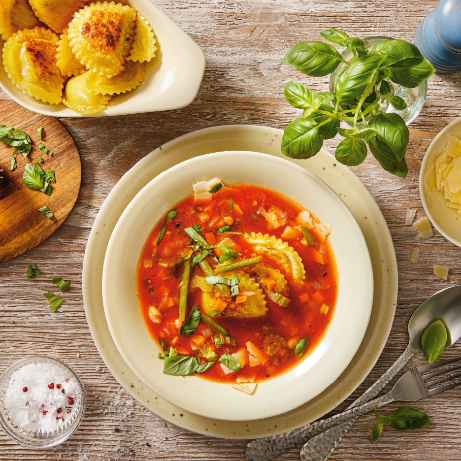 Ravioli i tomatsuppe med grøntsager, basilikum og parmesan på et træbord.