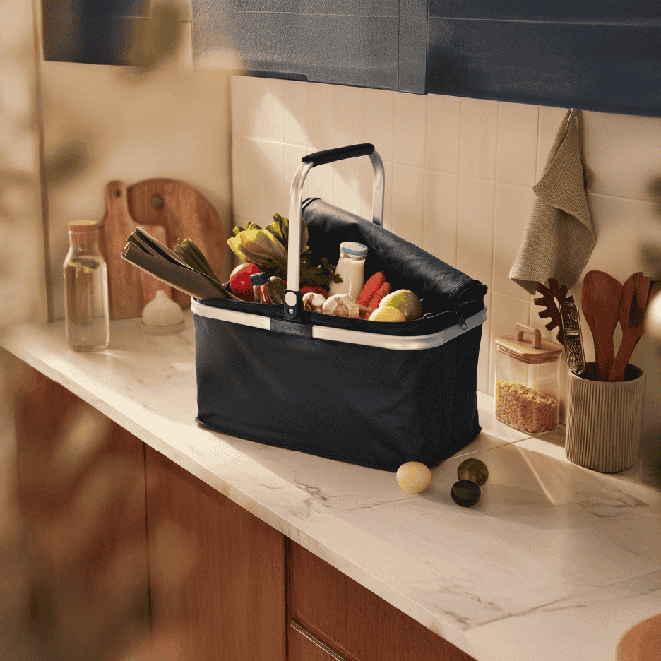 Indkøbskurv fyldt med friske grøntsager og dagligvarer på et køkkenbord.