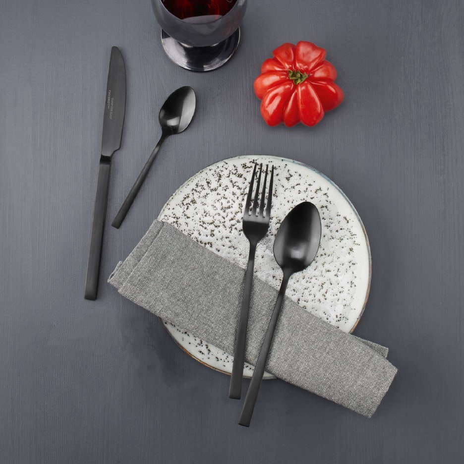 Sort bestik, grå serviet og hvid tallerken på et mørkegråt bord med en rød tomat.
