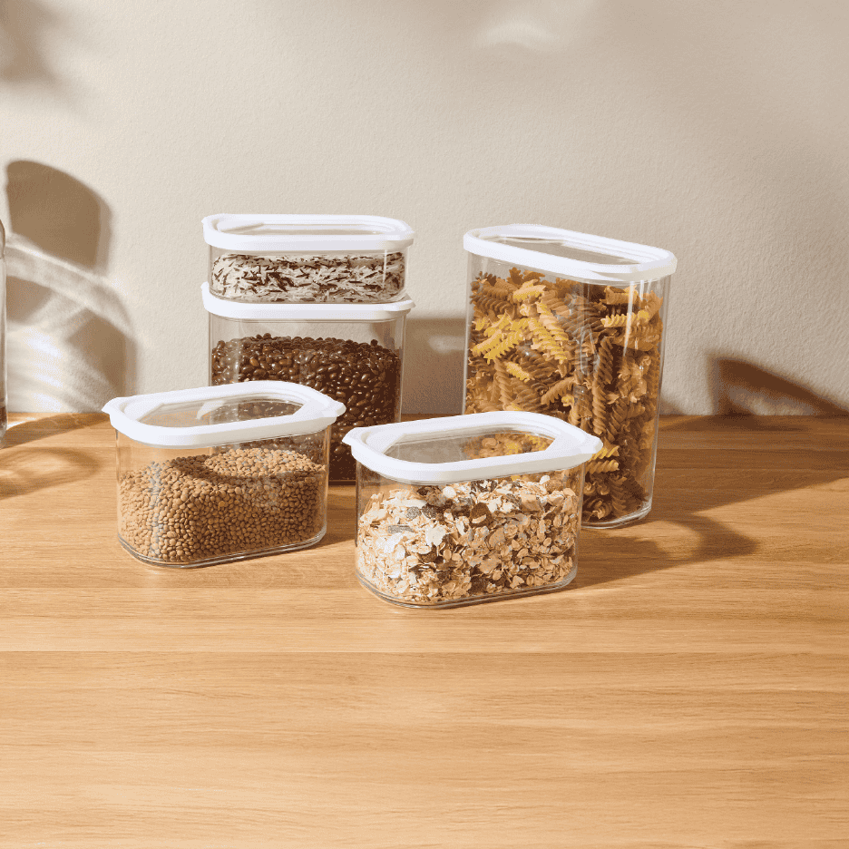 Gennemsigtige opbevaringsbeholdere fyldt med linser, havregryn, kaffe, ris og pasta på et træbord.