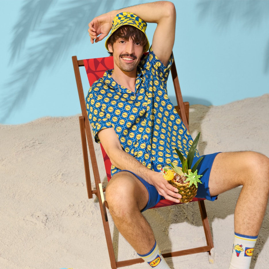 Mand i matchende bucket hat og skjorte med logo, blå shorts og sokker, der sidder i en strandstol med en ananasdrink.