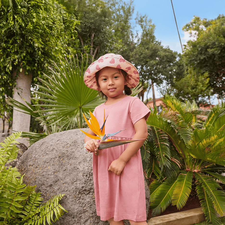 Lille pige i lyserød kjole og solhat holder en paradisfuglblomst i en frodig have.