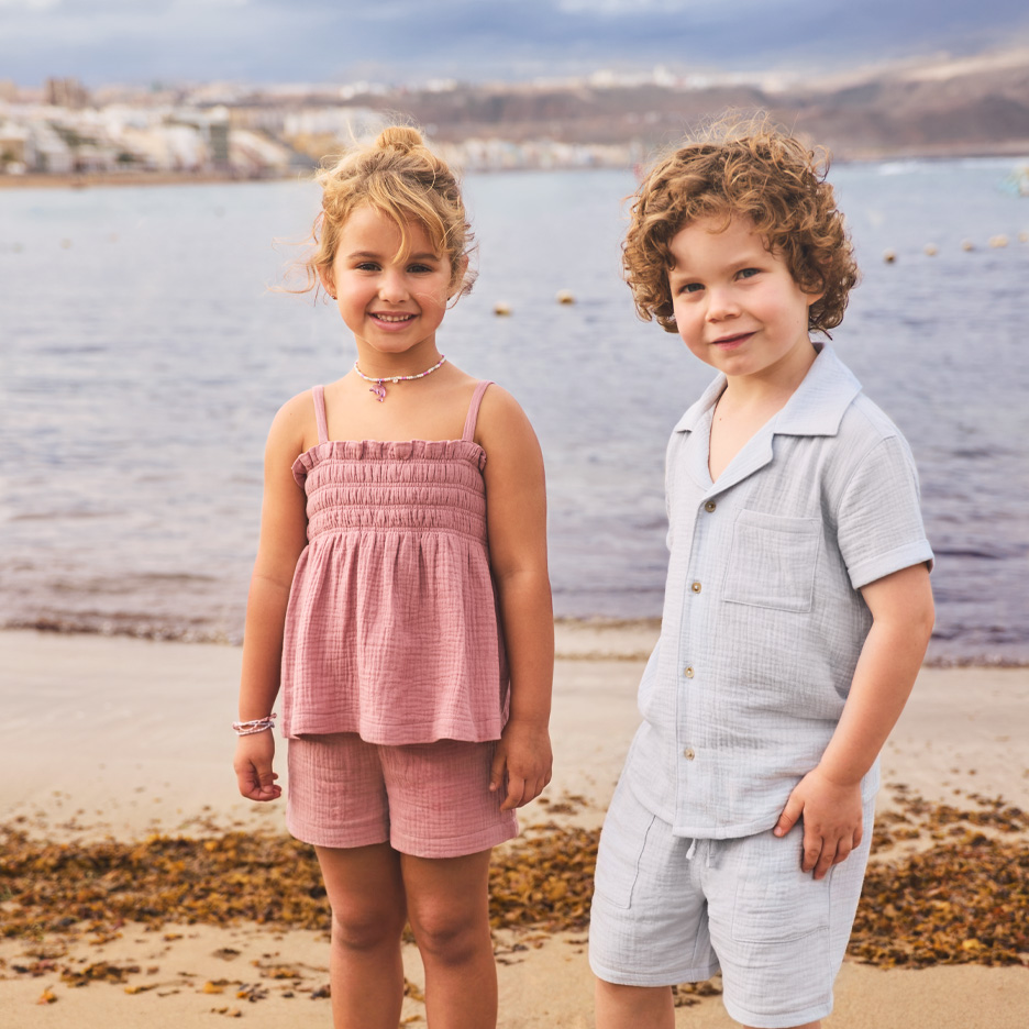 To børn i matchende sommeroutfits på stranden, en pige i lyserød og en dreng i lyseblå.