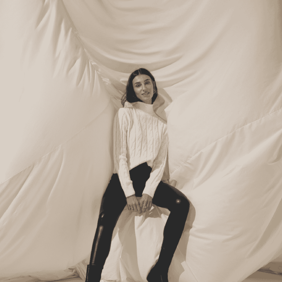 Kvinde i hvid kabelstrikket sweater og sorte bukser sidder foran et hvidt klæde.