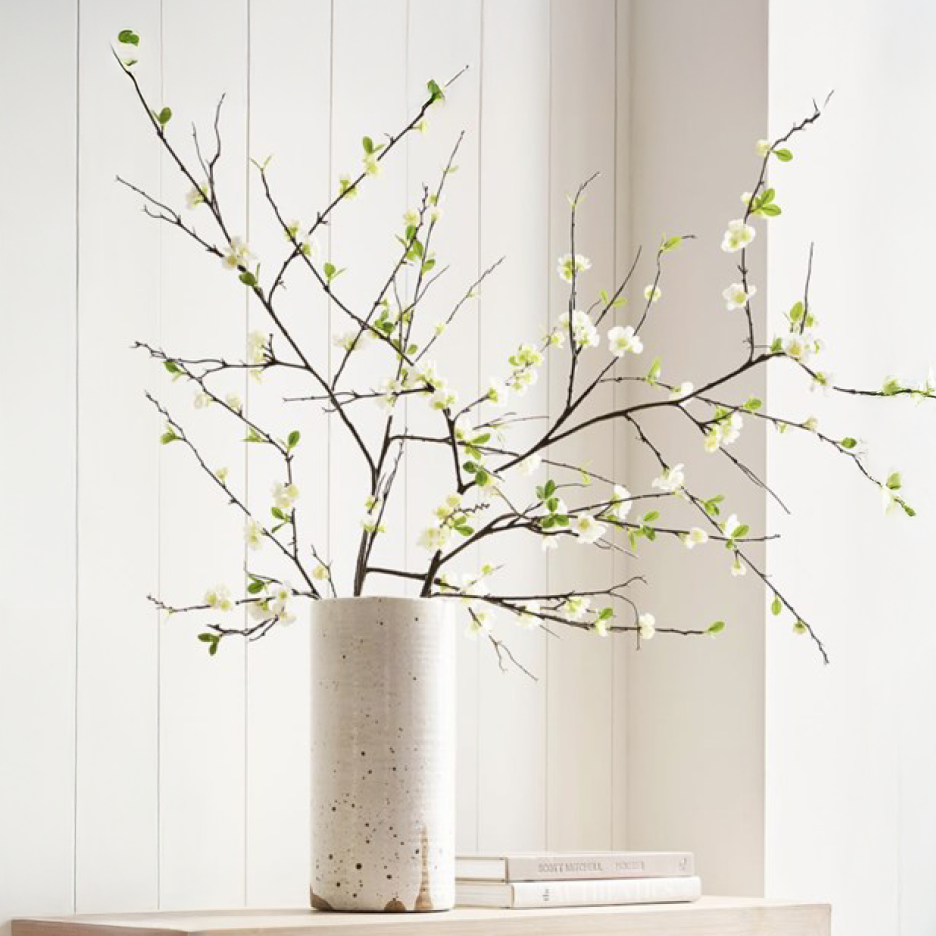 Hvide blomstrende grene i en høj, plettet vase på en træhylde med bøger.