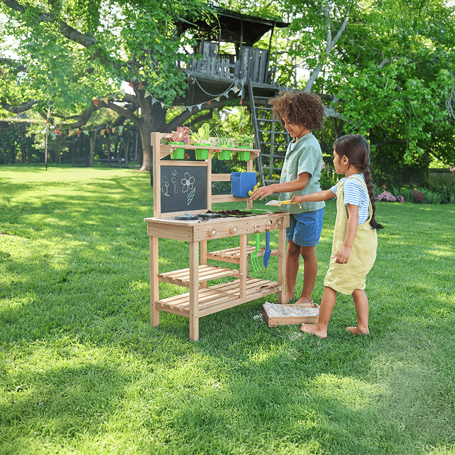To børn leger med et træhavebord med planter og redskaber i en grøn have.