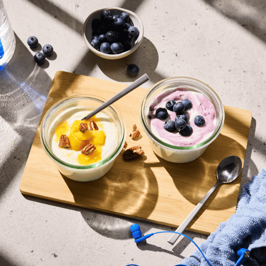 To skåle yoghurt med frugt og nødder på et træskærebræt, med blåbær og hovedtelefoner.