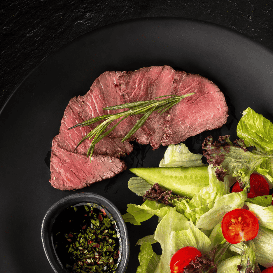 Skiver af oksekød med rosmarin, salat og dressing på en sort tallerken.