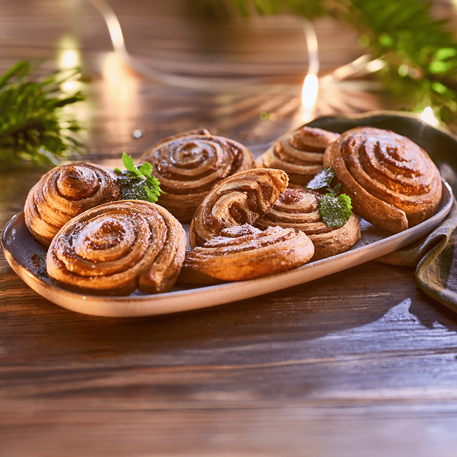 Kanelsnegle med flormelis og mynte på et fad, omgivet af julelys og grangrene.