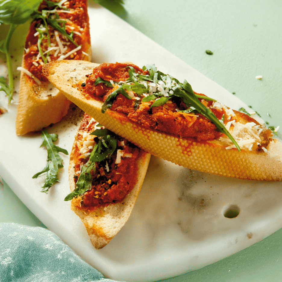 Bruschetta med tomatsauce, rucola og parmesanost på et hvidt marmorbræt.