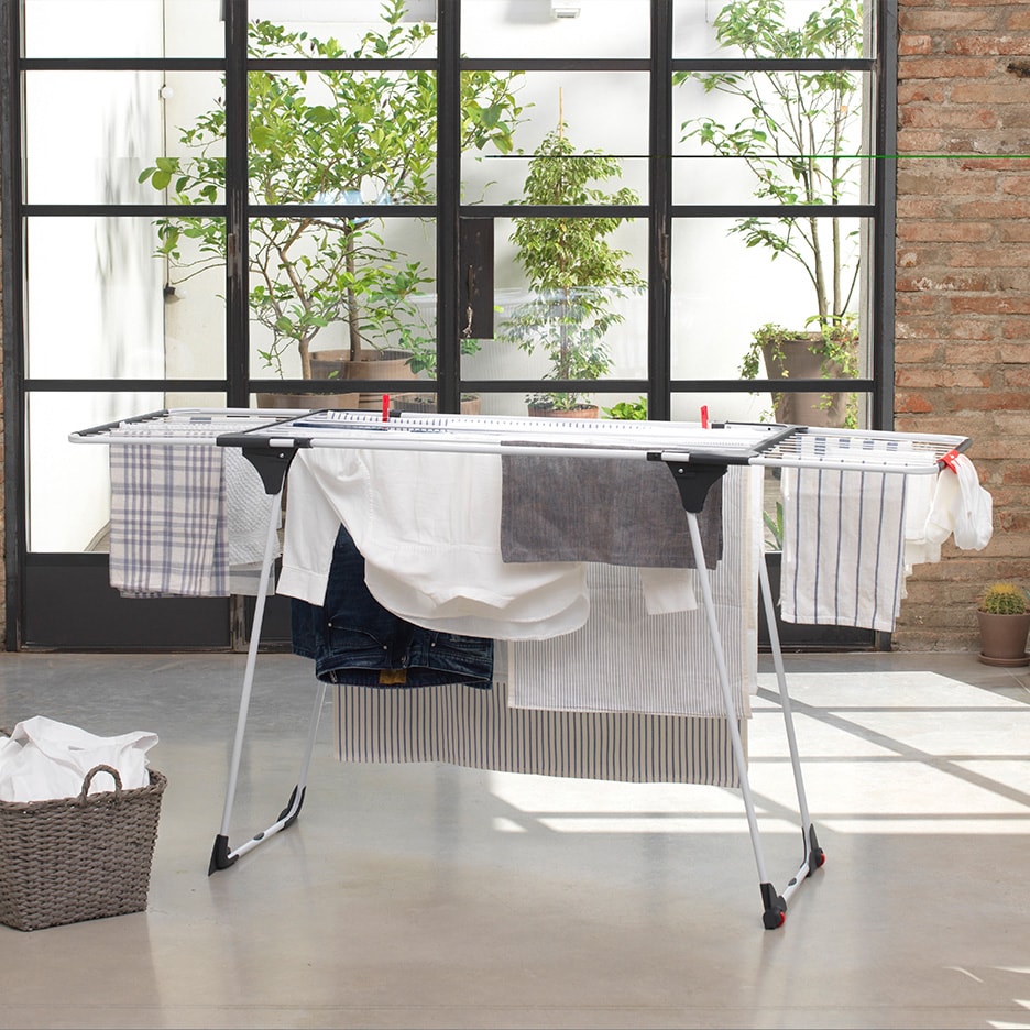 Tøjstativ med tøj, der tørrer, foran et stort vindue med planter.