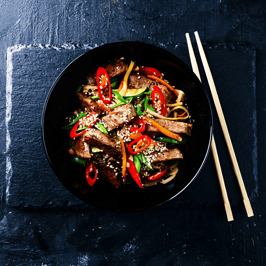 Oksekødswok med grøntsager, nudler, chili og sesamfrø i en sort skål med spisepinde.