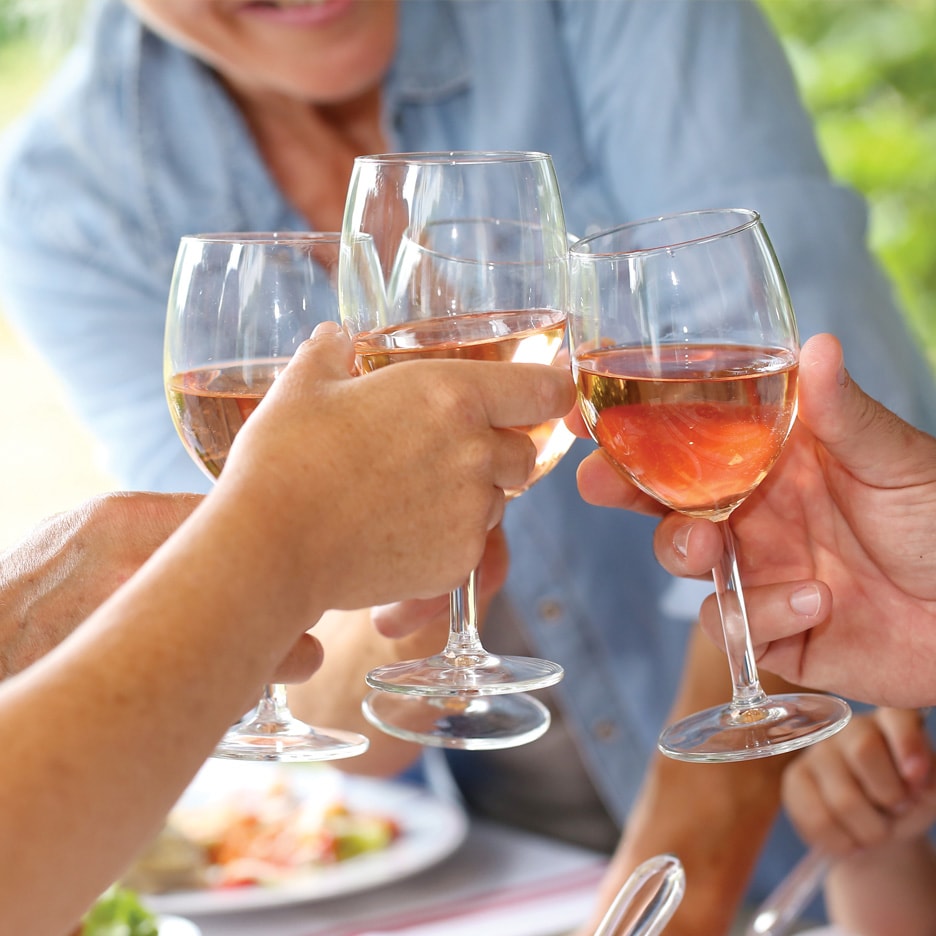 Fire personer skåler med glas fyldt med rosévin ved et udendørs bord.