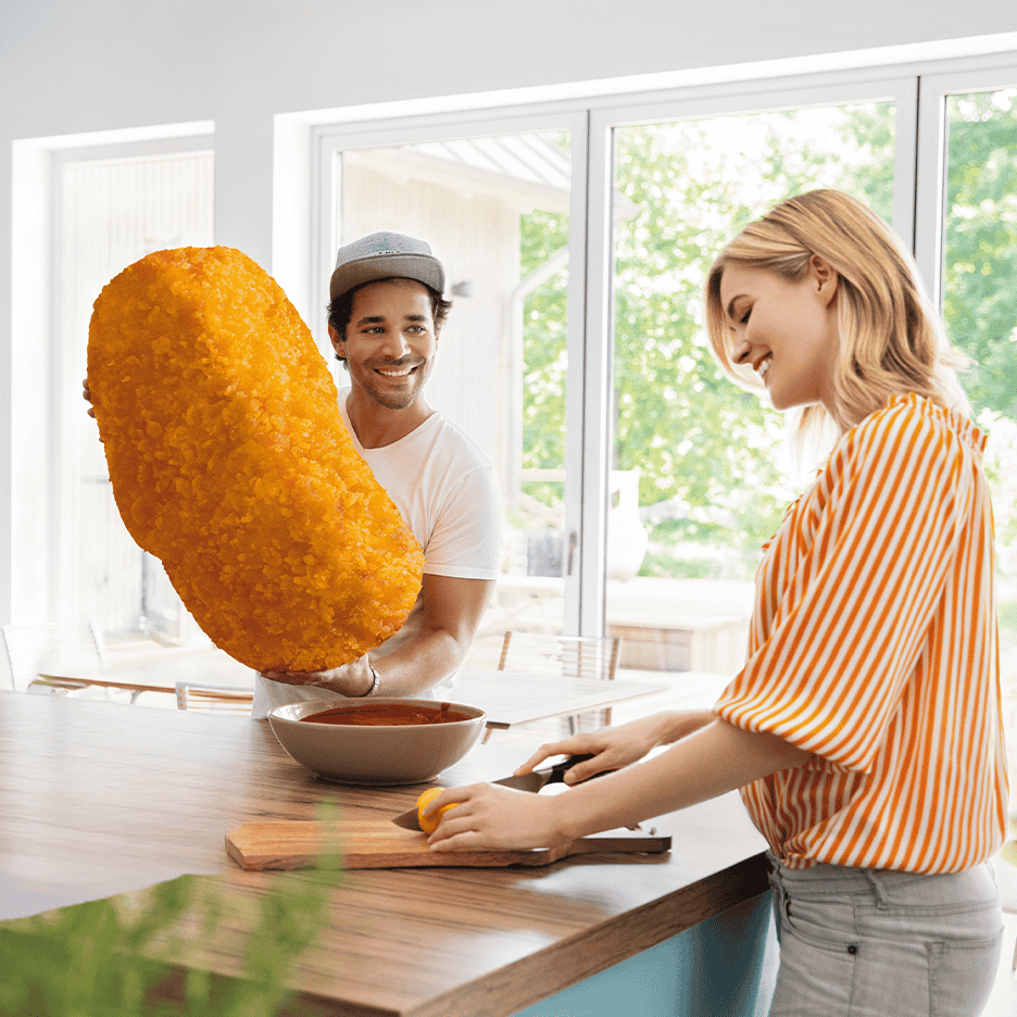 Mand holder kæmpe kroket, kvinde skærer citron i et lyst køkken.