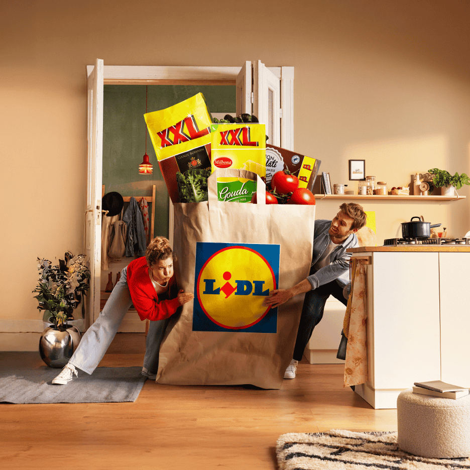 Par skubber en kæmpe Lidl-pose fyldt med XXL-produkter, Gouda-ost og tomater ind i et køkken.