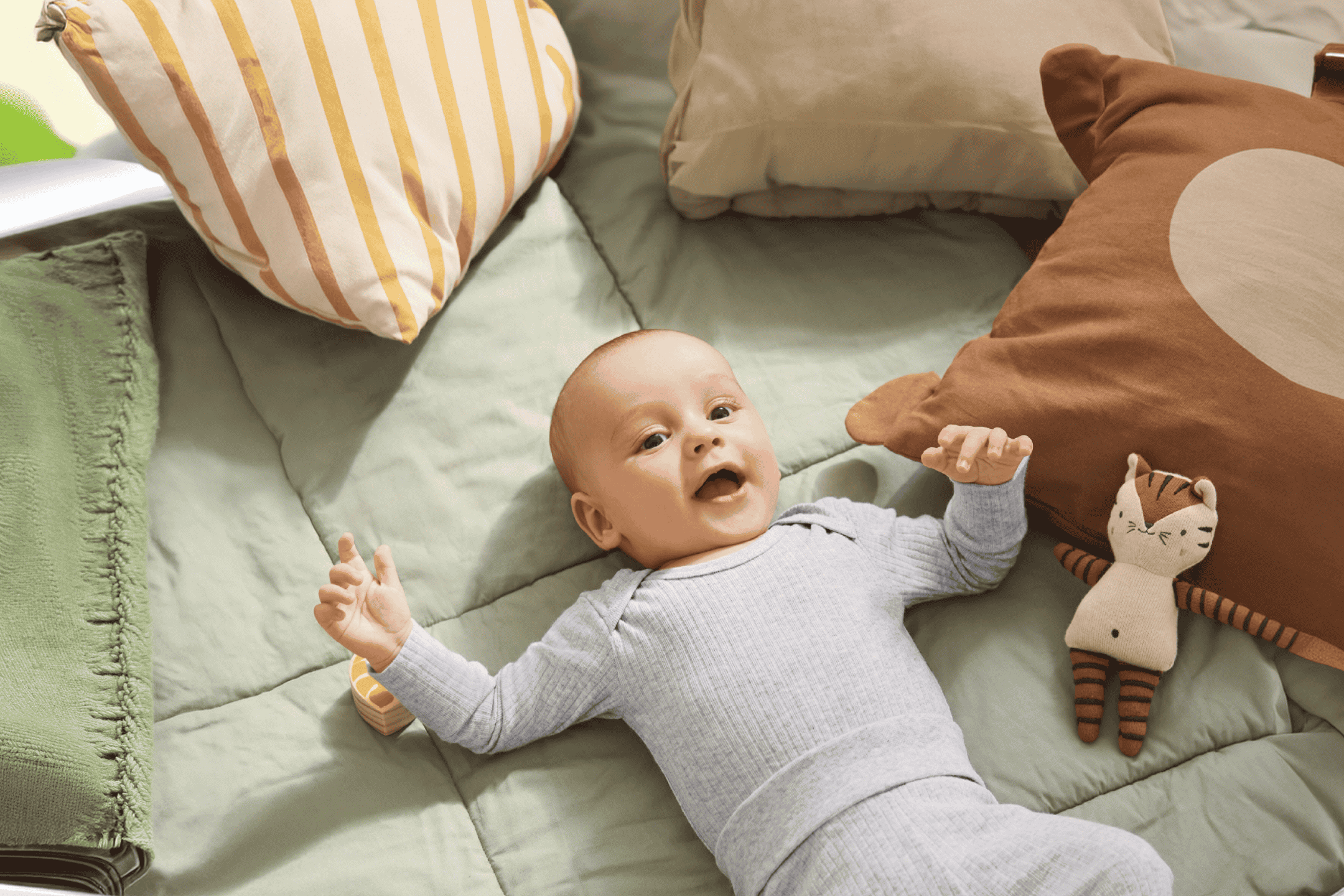 Glad baby i grå sparkedragt liggende på grønt tæppe omgivet af puder og legetøj.