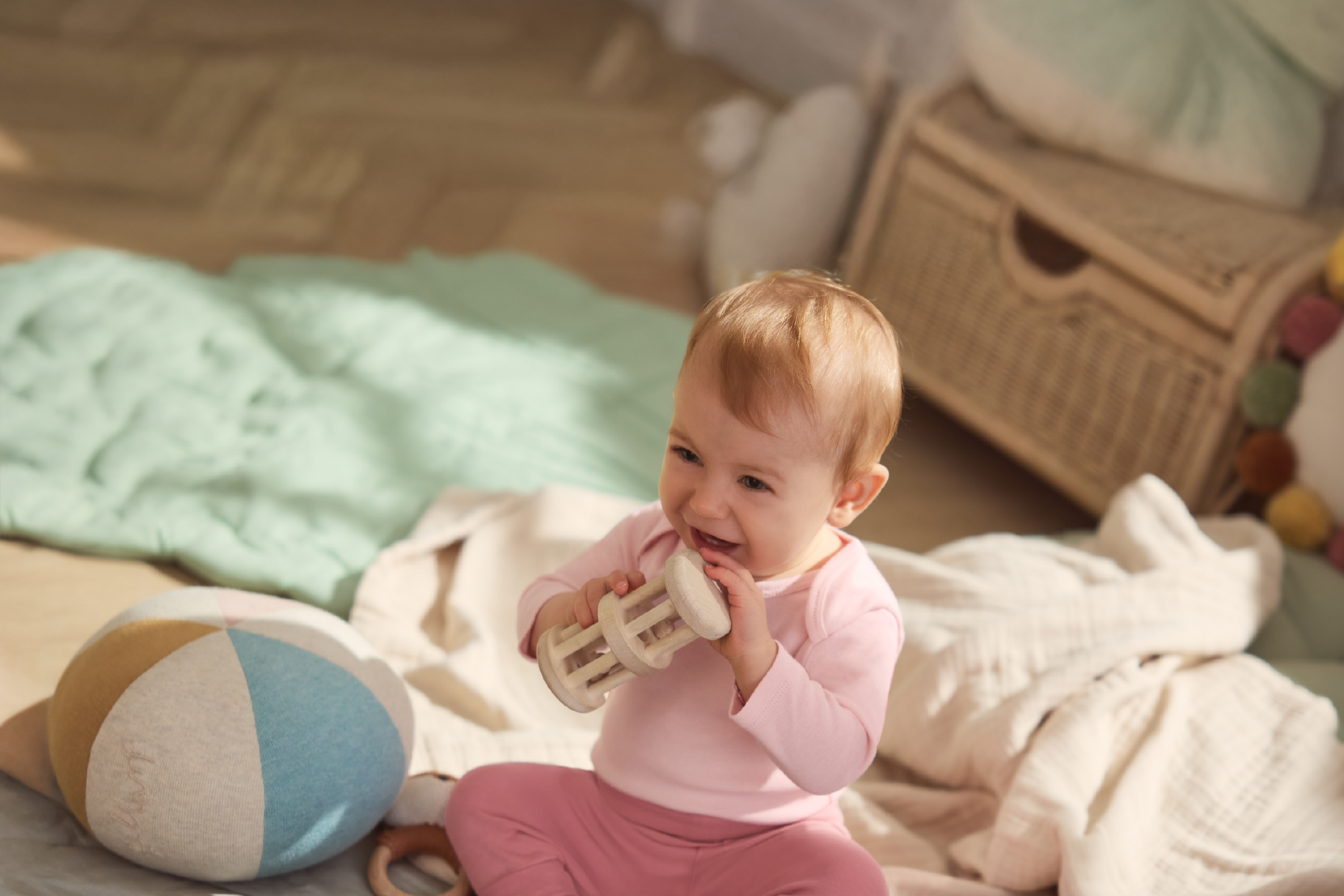 Glad baby i lyserødt tøj leger med trærangle og farverig bold på et tæppe.