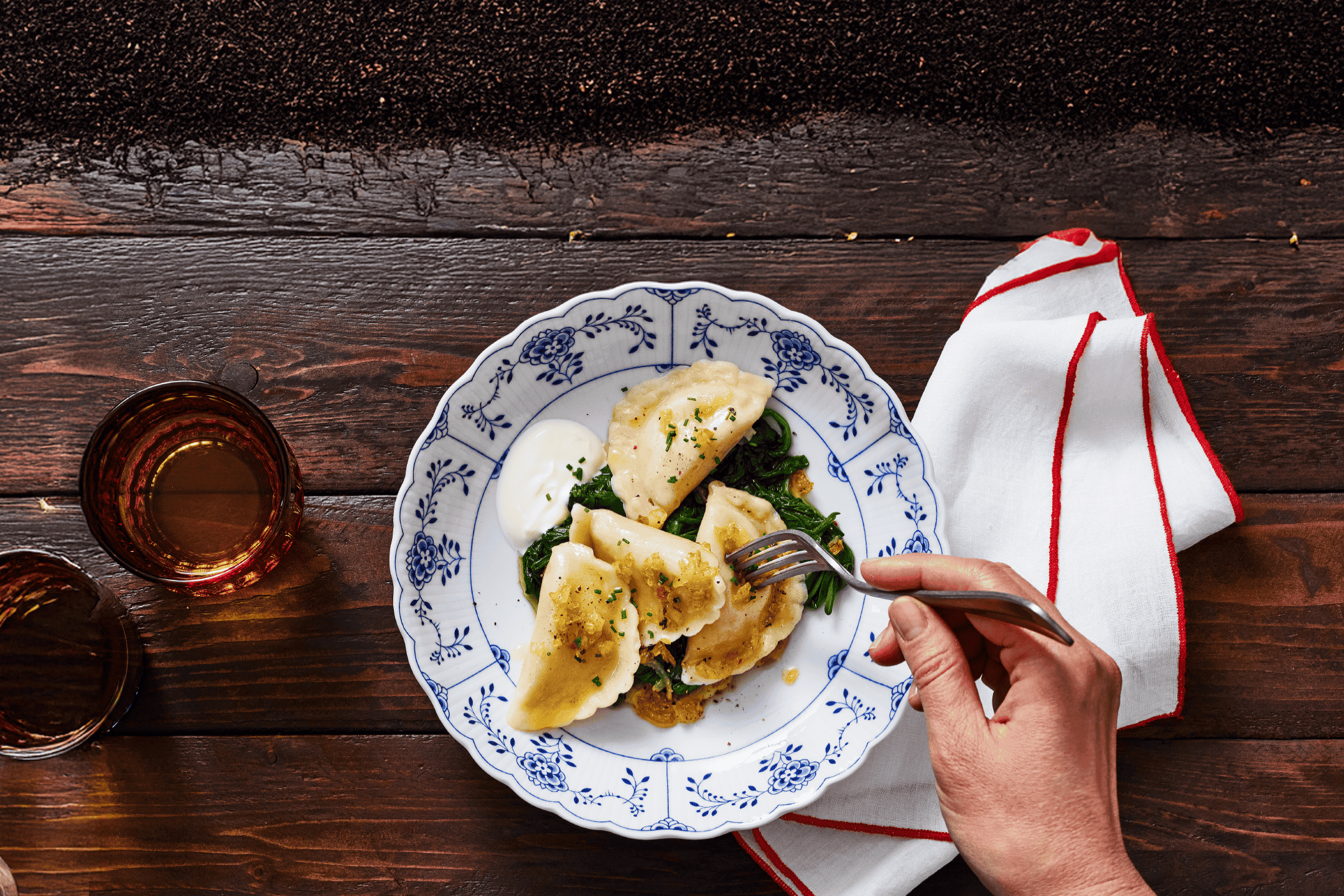 Hånd der tager en pierogi fra en tallerken med pierogi, spinat og creme fraiche, med to glas drikkevarer.
