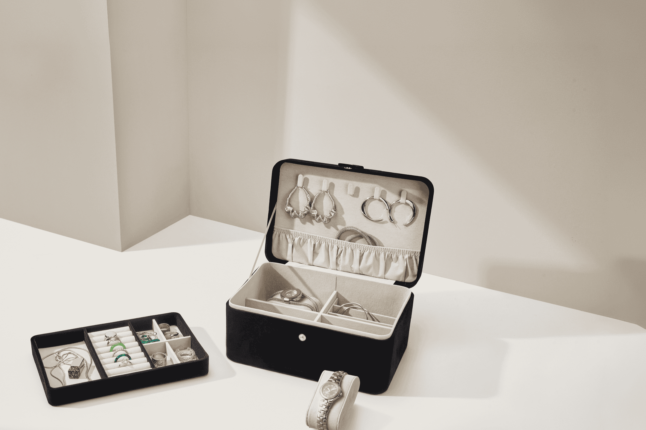 Åben sort smykkeskrin med øreringe, ure og ringe, samt en sort smykkebakke.