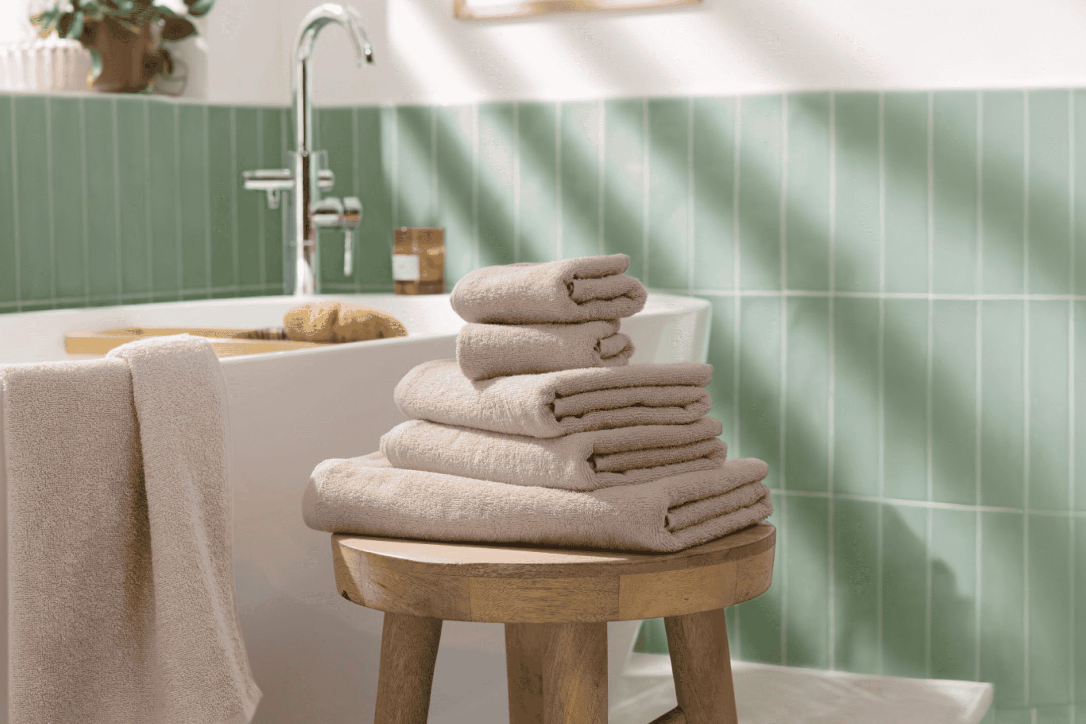 Stabel af beige håndklæder på en træskammel i et badeværelse med grønne fliser.