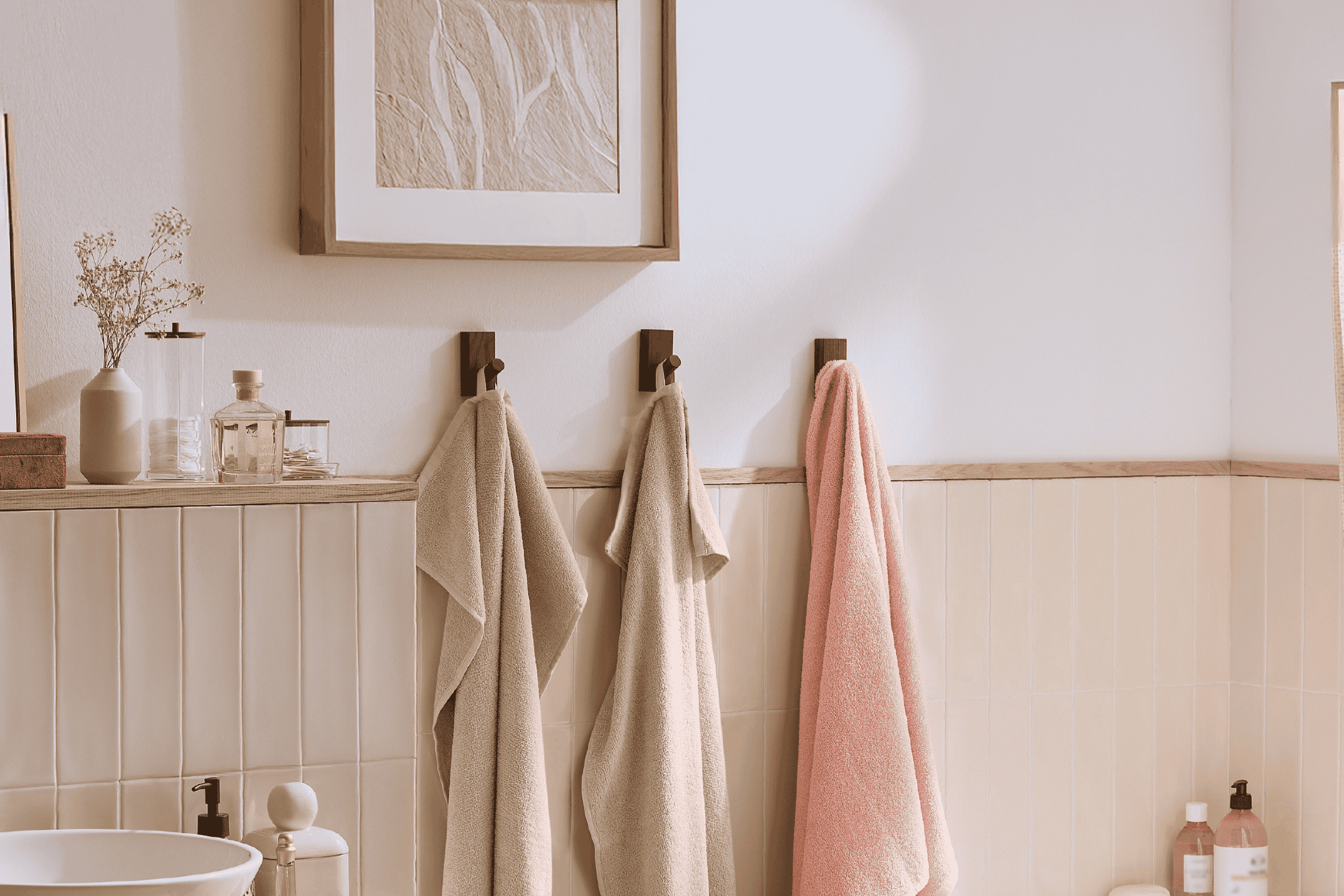 Badeværelse med tre håndklæder på kroge, en vase med tørrede blomster og en indrammet illustration.