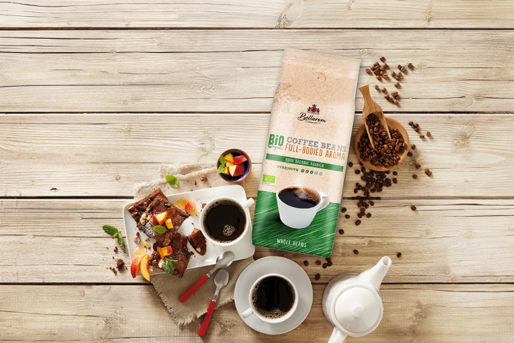 Kaffebønner i pose, kaffe i kopper, kage og frugt på et træbord.