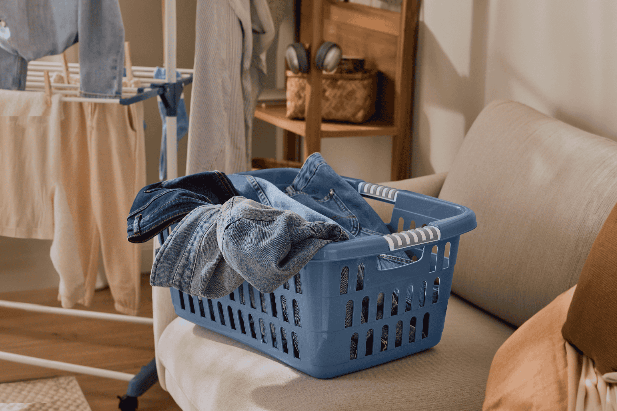 Vasketøjskurv fyldt med jeans på en sofa, med et tørrestativ i baggrunden.