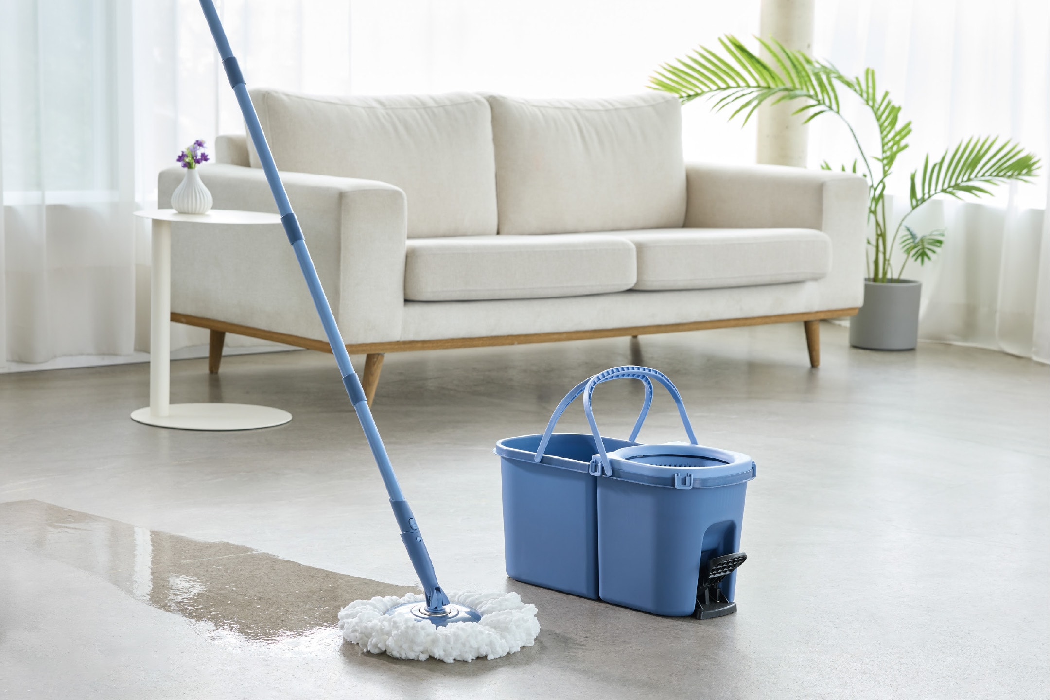 Blå moppe og spand med vand på gulvet i en stue med en sofa og plante.