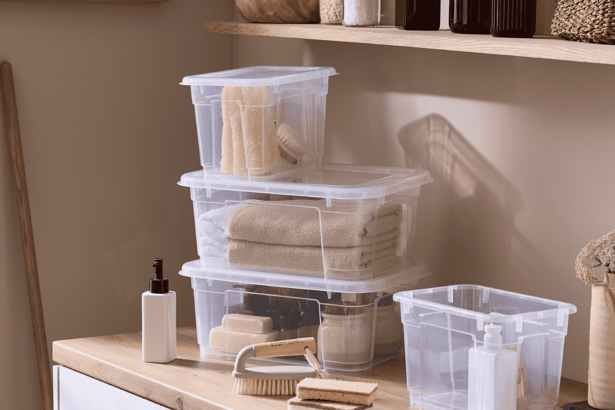 Stabelbare gennemsigtige opbevaringskasser med håndklæder og badeværelsesartikler på en træbordplade.