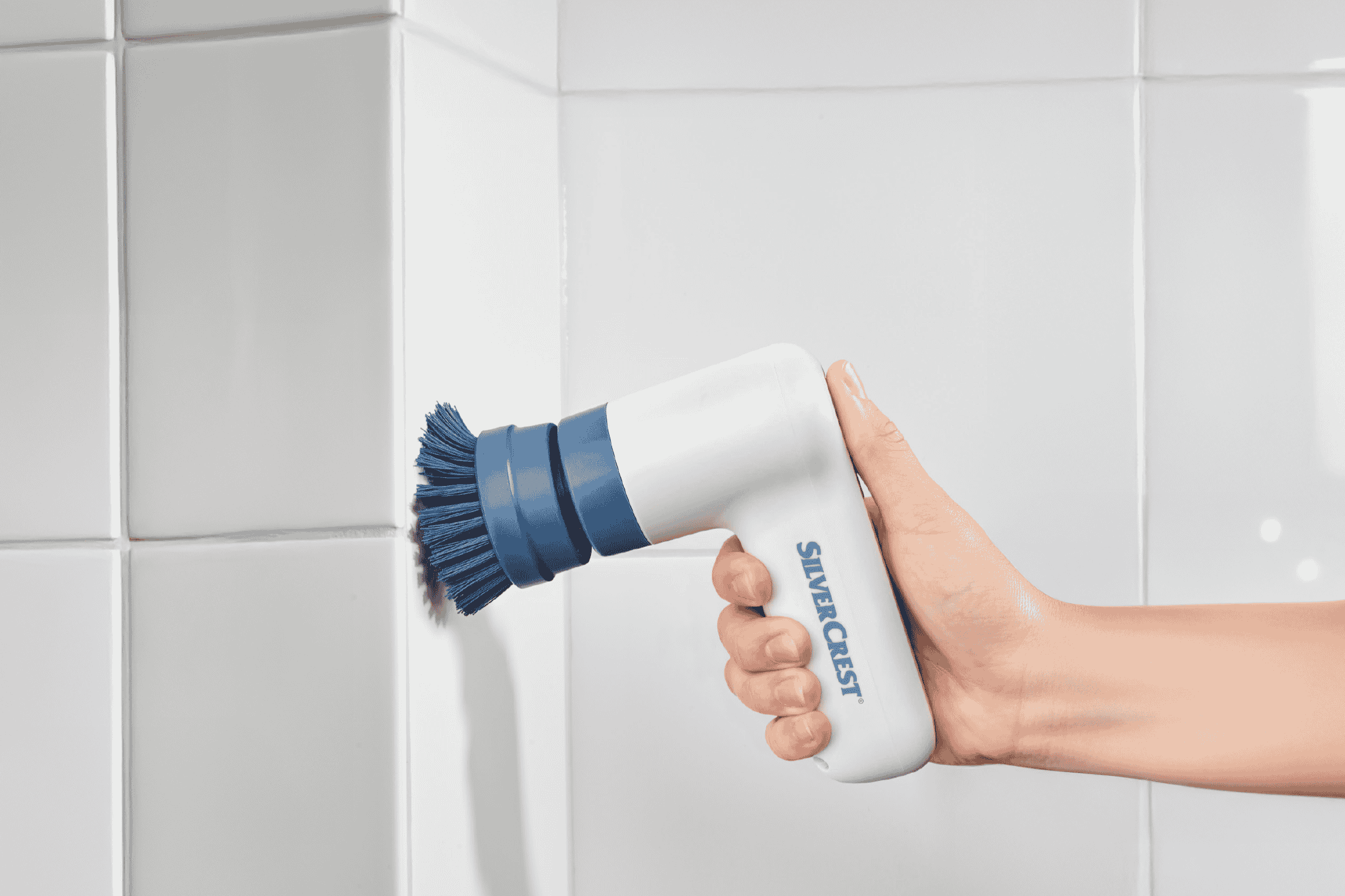 Hånd holder en hvid og blå elektrisk skrubbebørste mod hvide fliser.