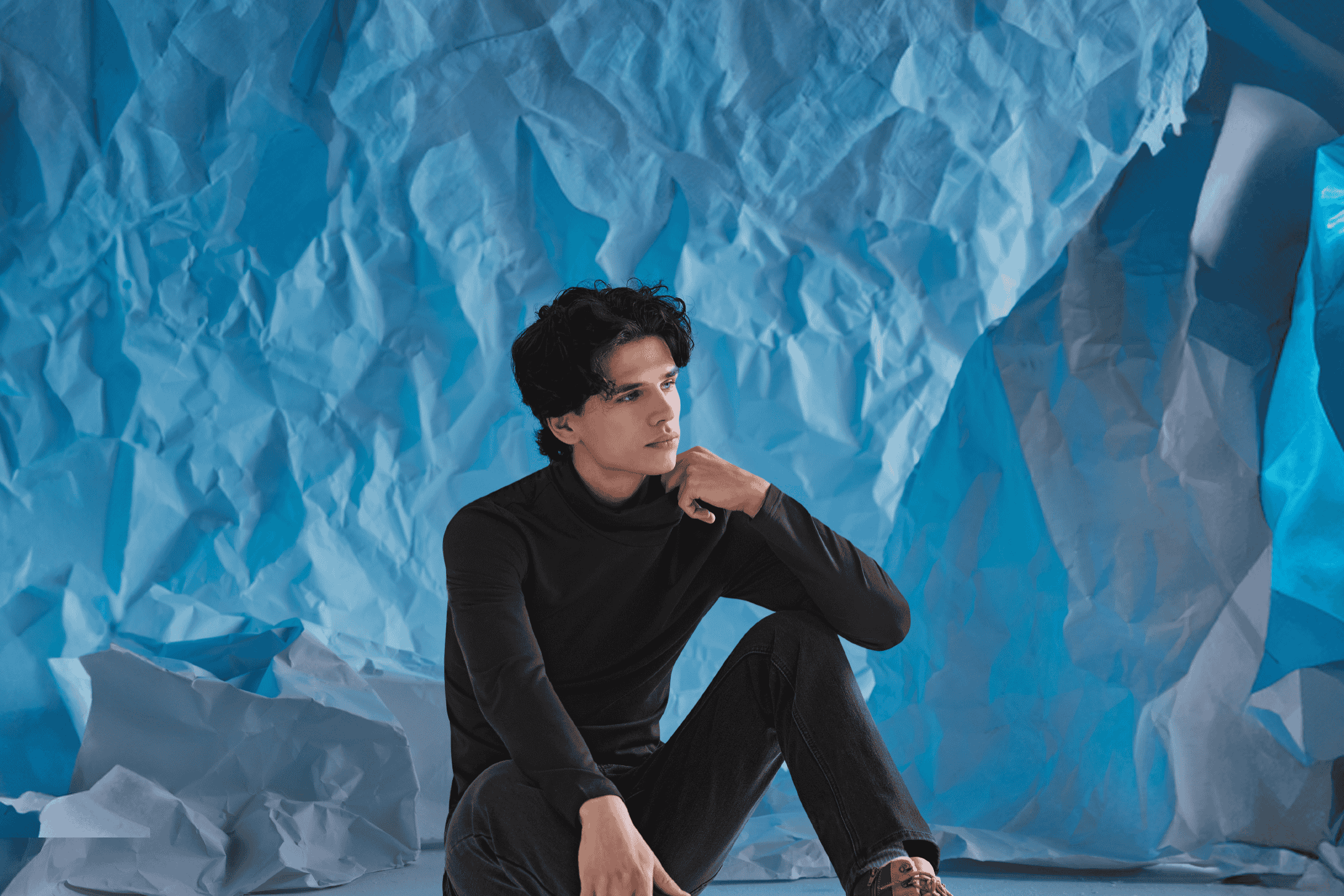 Mand i sort rullekrave og jeans sidder foran en krøllet blå baggrund.