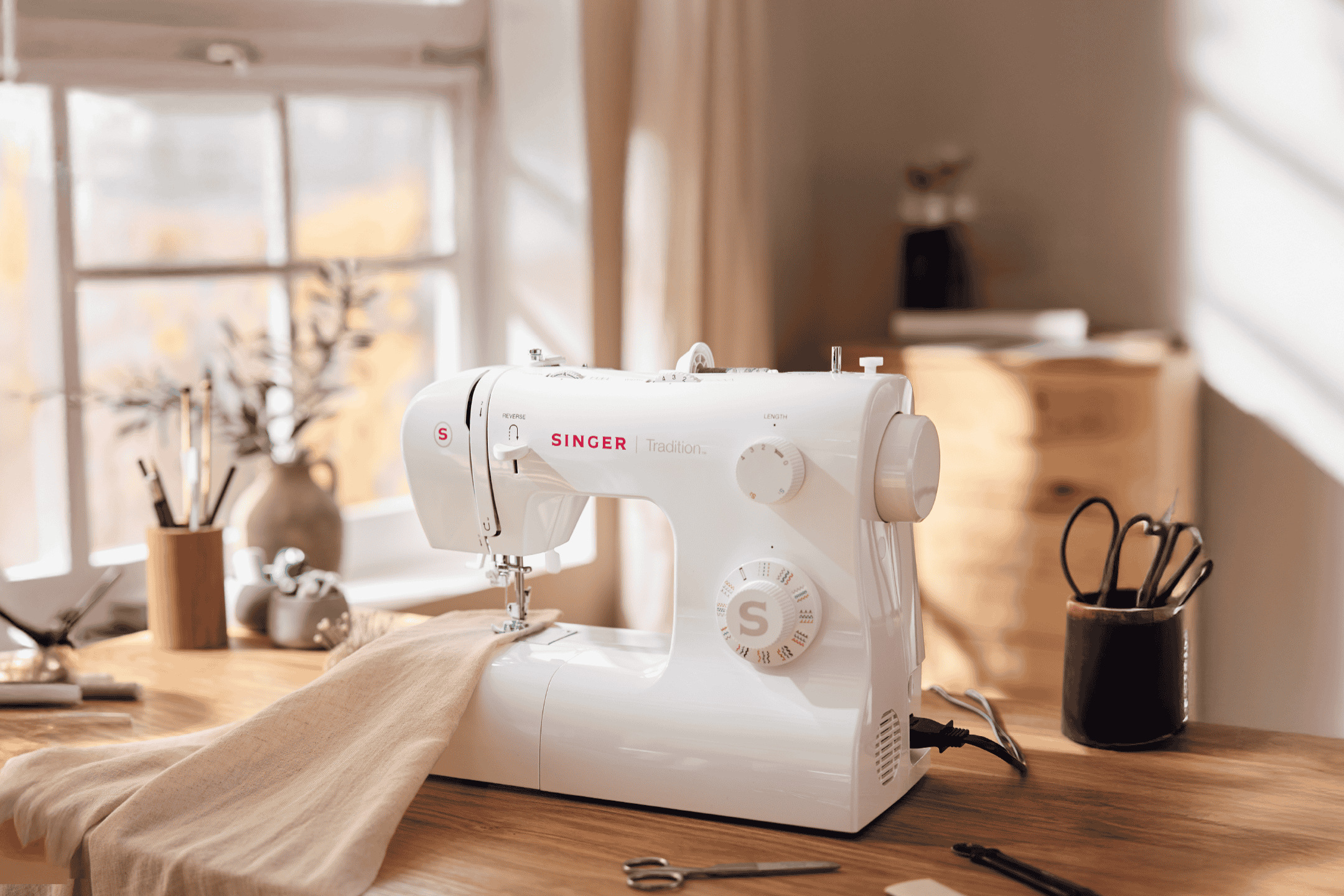 Symaskine på et træbord med stof og syartikler i et lyst rum.