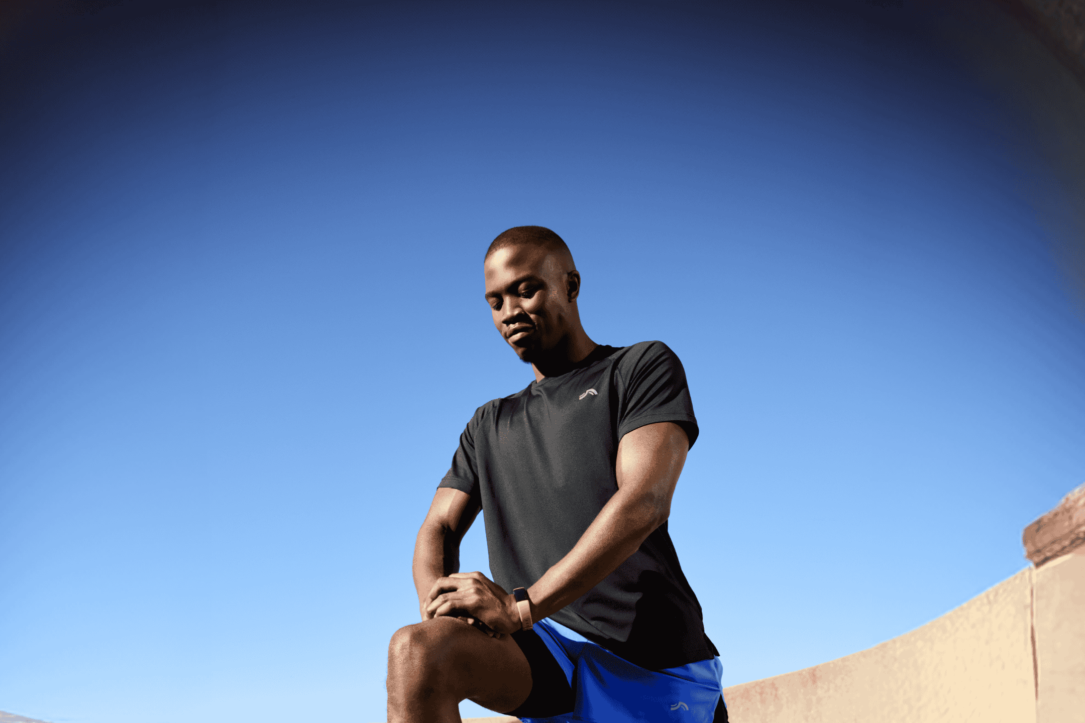 Mand i sort trænings-T-shirt og blå shorts strækker sig under en klar blå himmel.