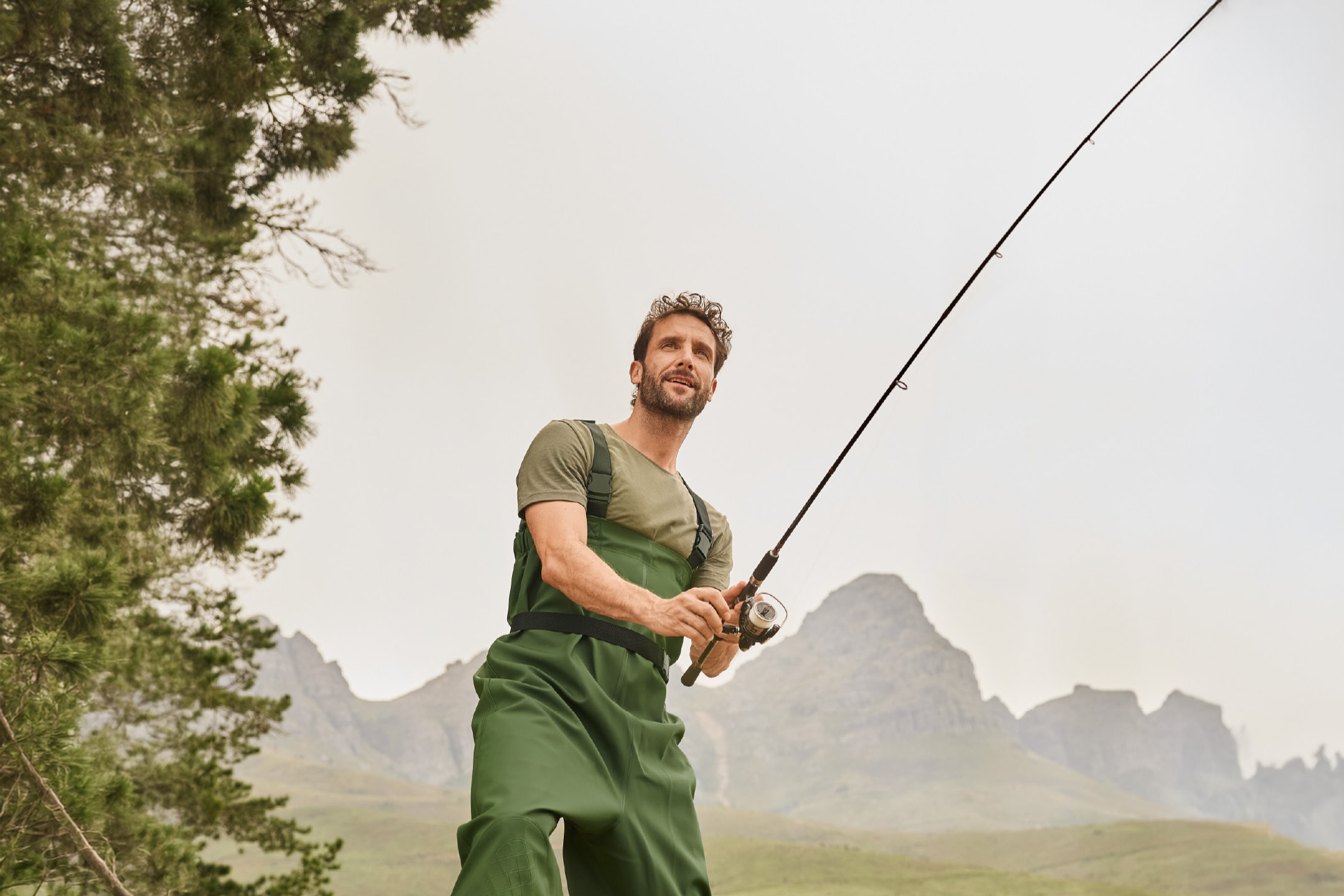 Mand i grønne waders og t-shirt fisker med fiskestang i bjerglandskab.